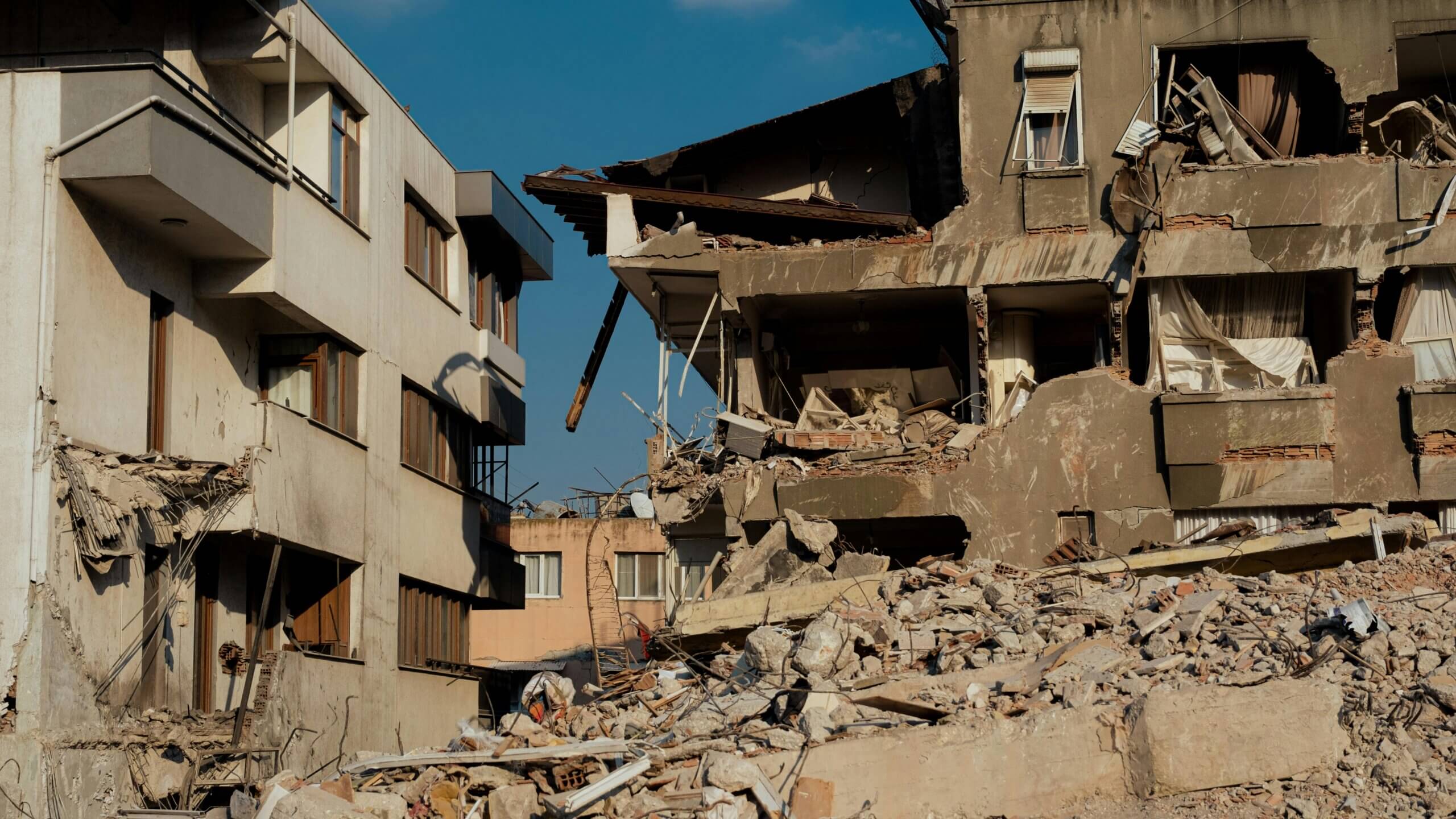 Partially collapsed residential buildings with rubble and debris under a clear blue sky.