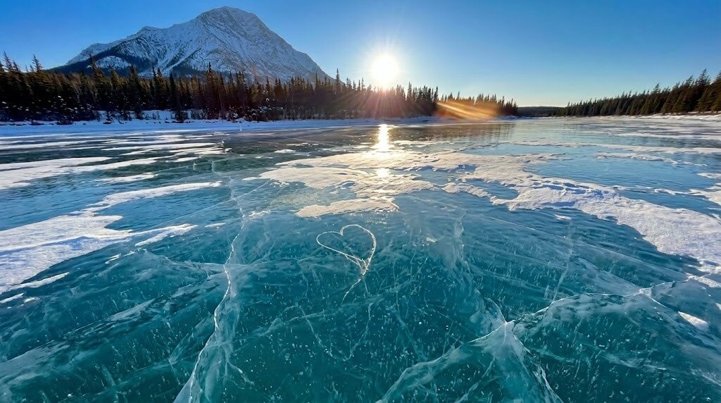 Frozen lake with a heart shape etched in the ice, surrounded by snow-covered trees and a mountain at sunset.
