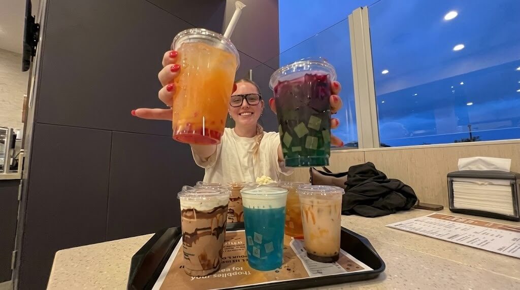 Person holding two colorful bubble tea drinks with six other bubble teas on a tray in a cafe.