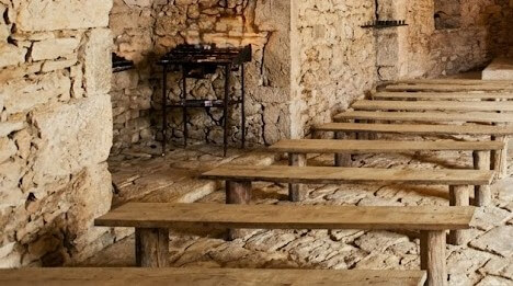 Rustic stone room with wooden benches and a metal grill against the wall.