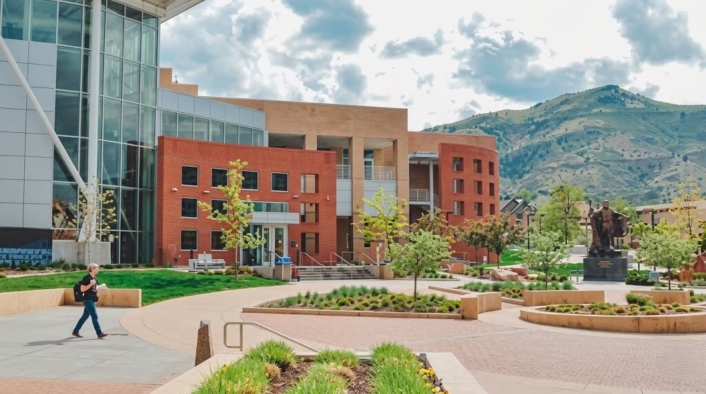 Student walking near modern university buildings with mountains in the background and a statue in the courtyard