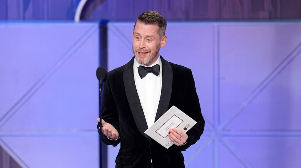 Man in black tuxedo holding an envelope speaking at a microphone on a purple-lit stage.
