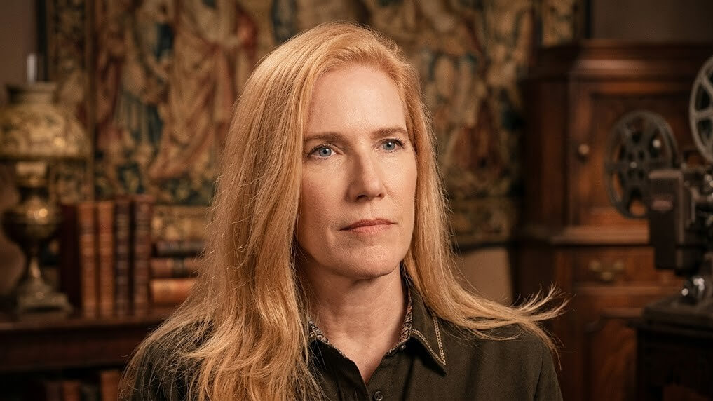 Blonde woman with long hair wearing a dark shirt in a room with vintage books and film equipment.