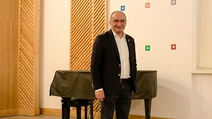 Man in black suit and white shirt standing in front of a covered piano in a room with wooden paneling and colorful square wall decorations
