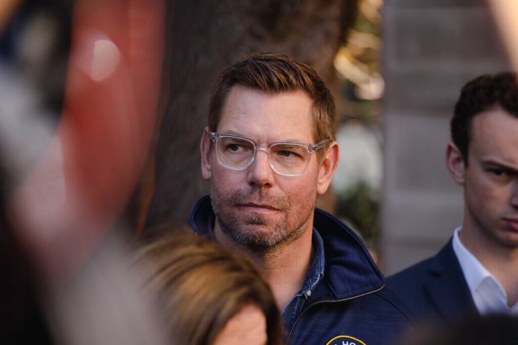 Man with clear glasses and blue jacket looking to the side in an outdoor setting with blurred people around