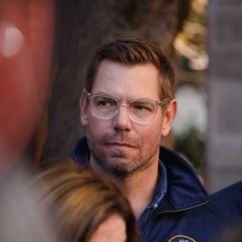 Man with clear glasses and blue jacket looking to the side in an outdoor setting with blurred people around