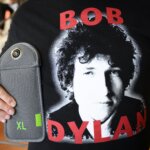 Person wearing a black Bob Dylan t-shirt holding a gray XL camera lens case.