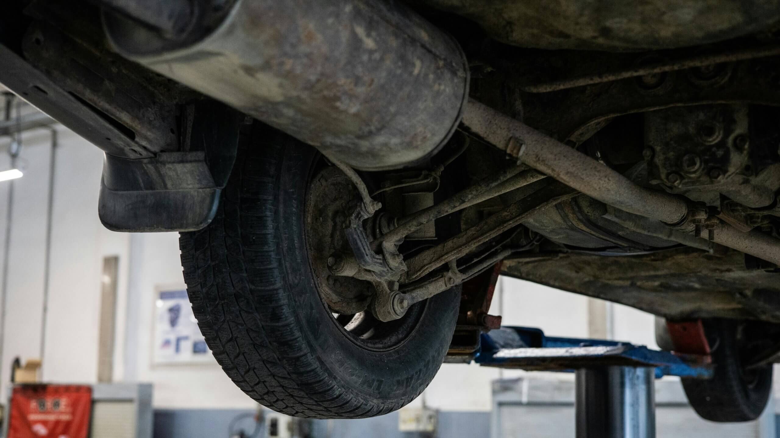 Underside of a vehicle showing rear suspension, exhaust system, and tire in an auto repair shop.