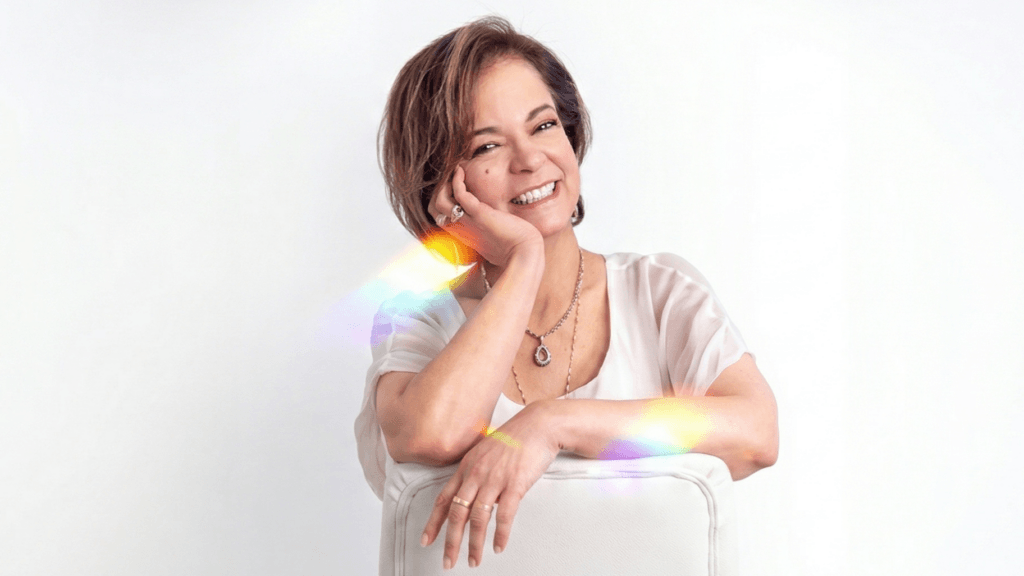 Smiling woman with short hair leaning on a white chair, wearing a white blouse and gold jewelry with rainbow light reflections.