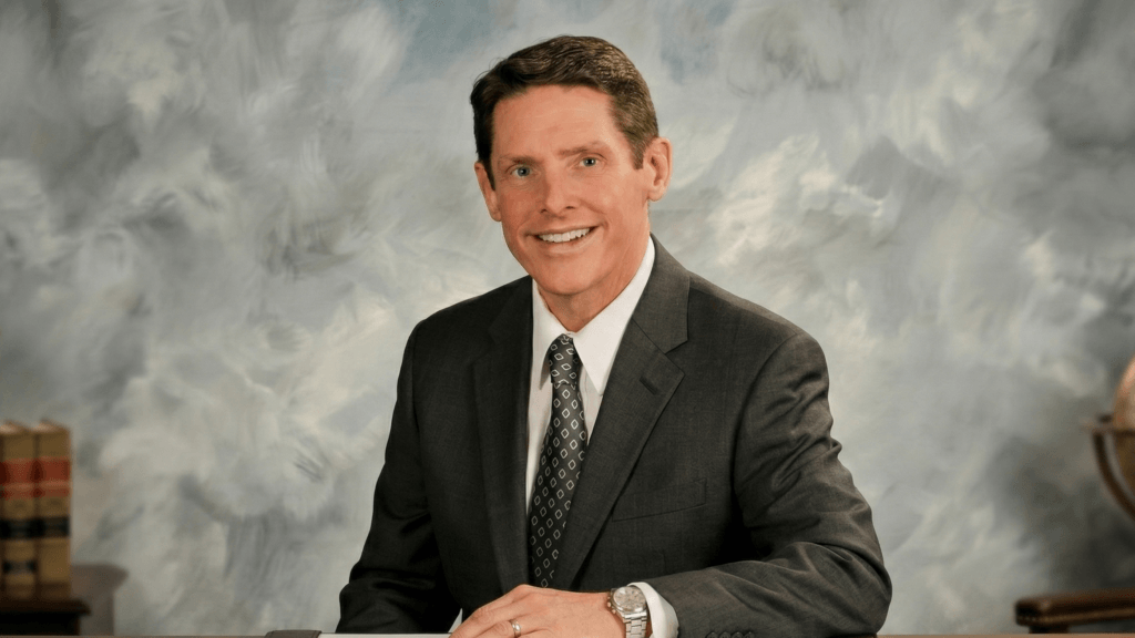 Smiling man in dark suit and patterned tie sitting at desk with books and globe in background