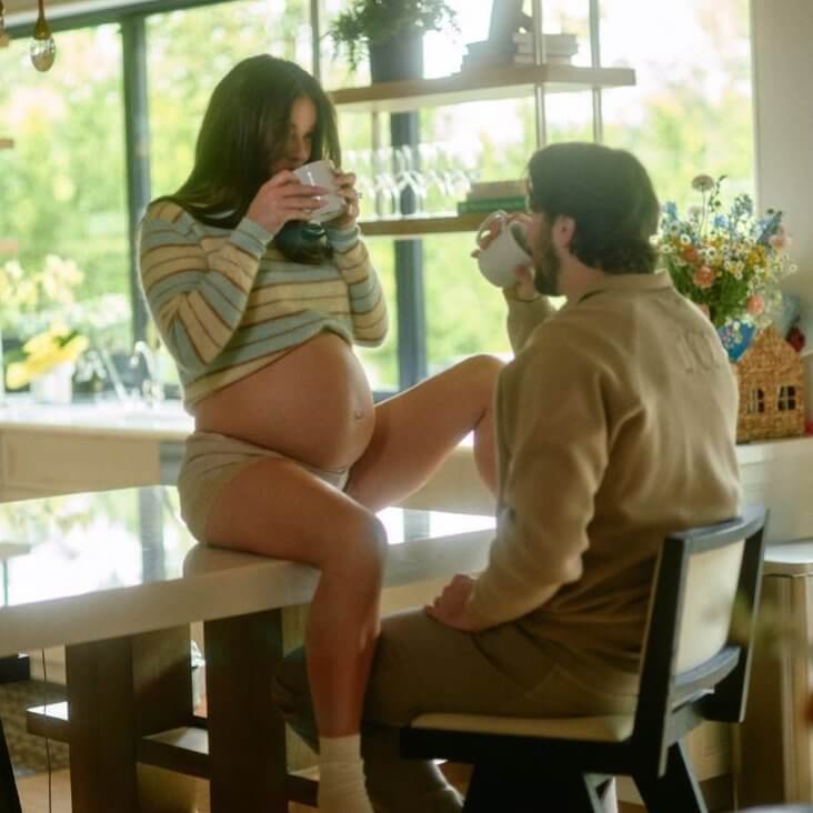 Pregnant woman in striped sweater and shorts drinking coffee with man in beige sweater in bright kitchen.