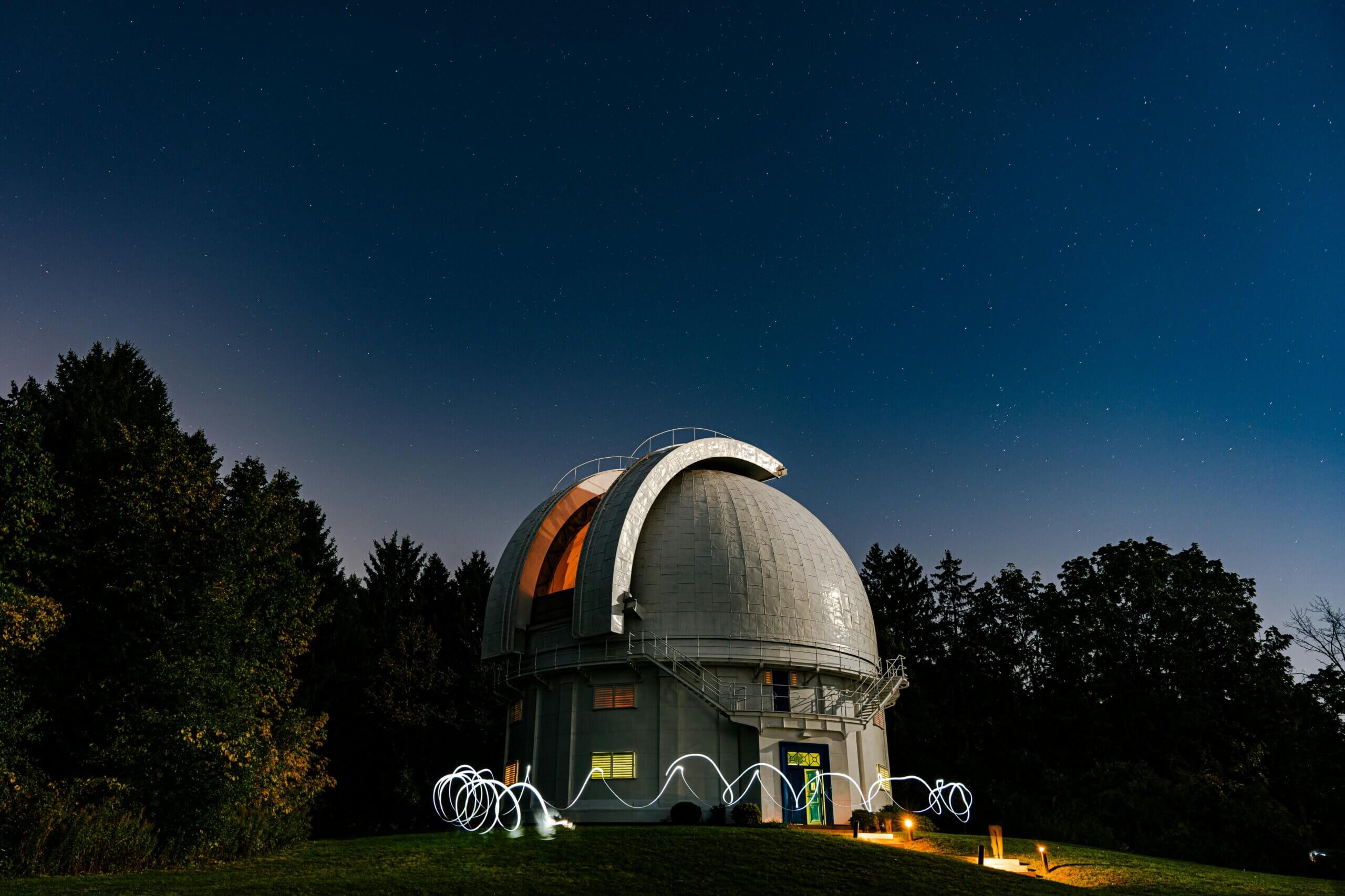 Observatory dome under a starry night sky with light trails on the grassy hill in front.