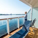 Woman in striped shirt standing on a cruise ship balcony overlooking calm water and distant port cranes.