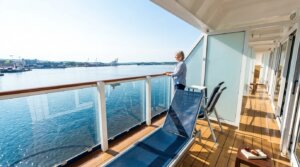 Woman in striped shirt standing on a cruise ship balcony overlooking calm water and distant port cranes.