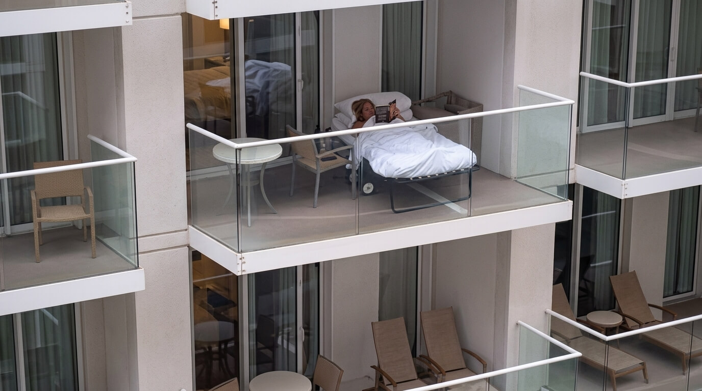 Person lying on a balcony bed covered with a white blanket, reading a book.