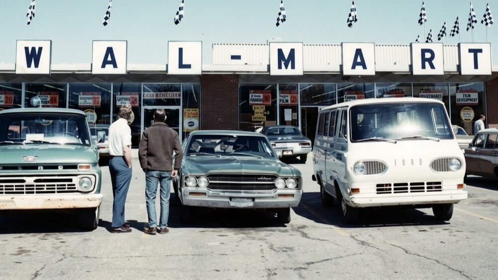 Two men stand in front of vintage cars parked outside a Walmart store with checkered flags above the sign.