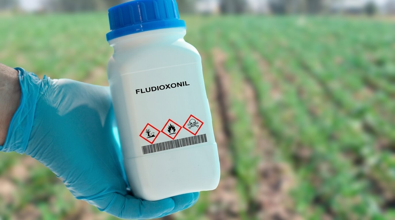 Hand in blue glove holding a bottle labeled Fludioxonil with hazard symbols in a field background