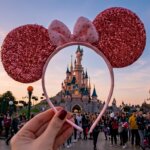Hand holding pink sequined Minnie Mouse ears headband framing Sleeping Beauty Castle at Disneyland with crowd in background.