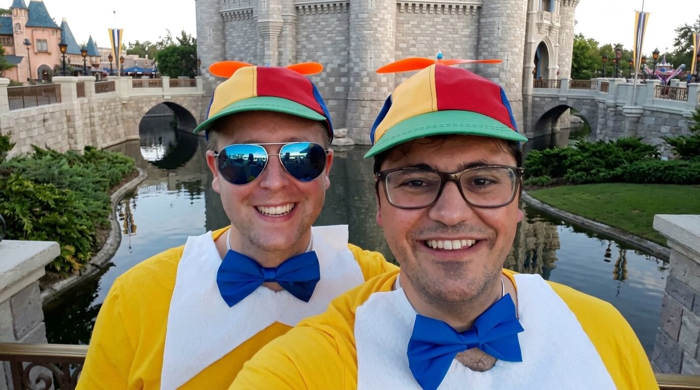 Two men smiling in colorful propeller hats and blue bow ties in front of a castle moat at a theme park