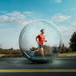 Man in red shirt running inside a transparent bubble on a road with trees and blue sky background.