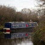 Narrowboat named The Hansebar cruising on a calm canal surrounded by leafless trees and bushes.