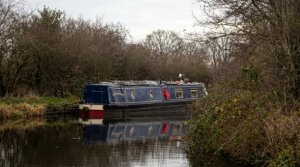 Narrowboat named The Hansebar cruising on a calm canal surrounded by leafless trees and bushes.
