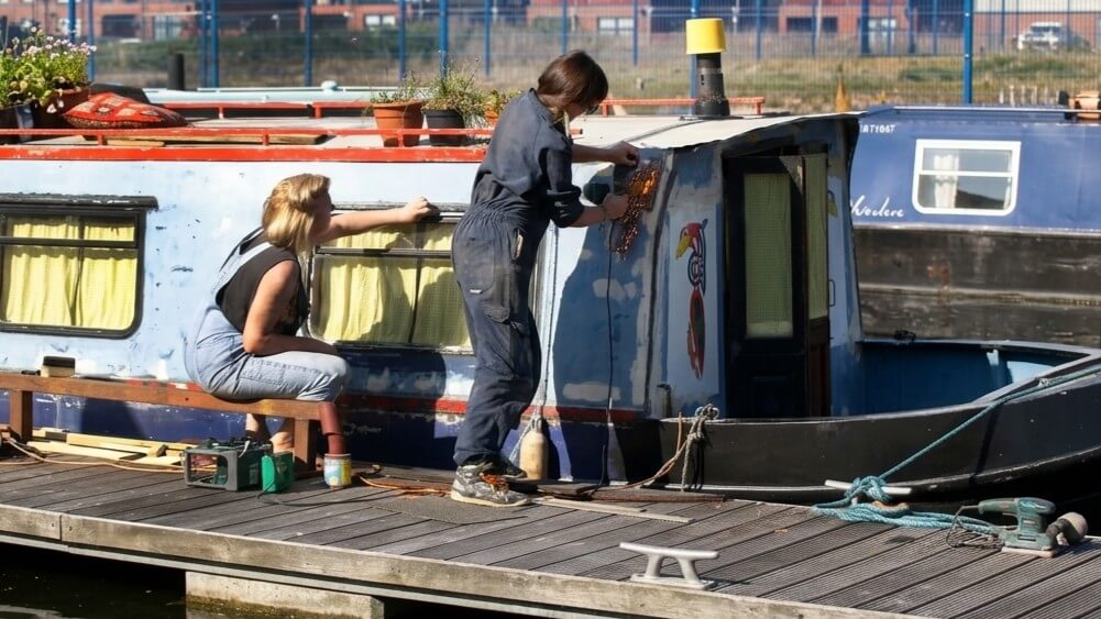 Two women working on the exterior of a narrowboat docked at a marina on a sunny day.