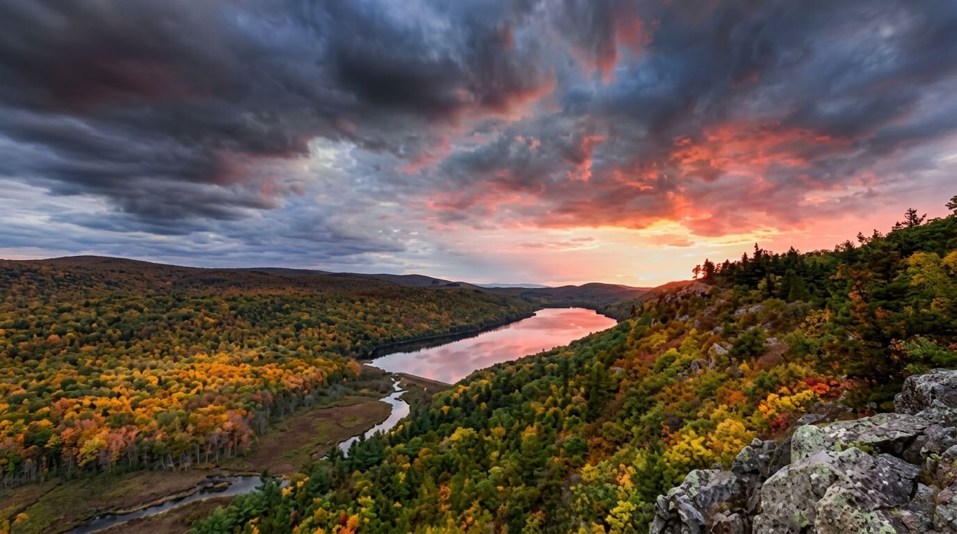 Sunset over a forested river valley with autumn foliage and dramatic clouds.