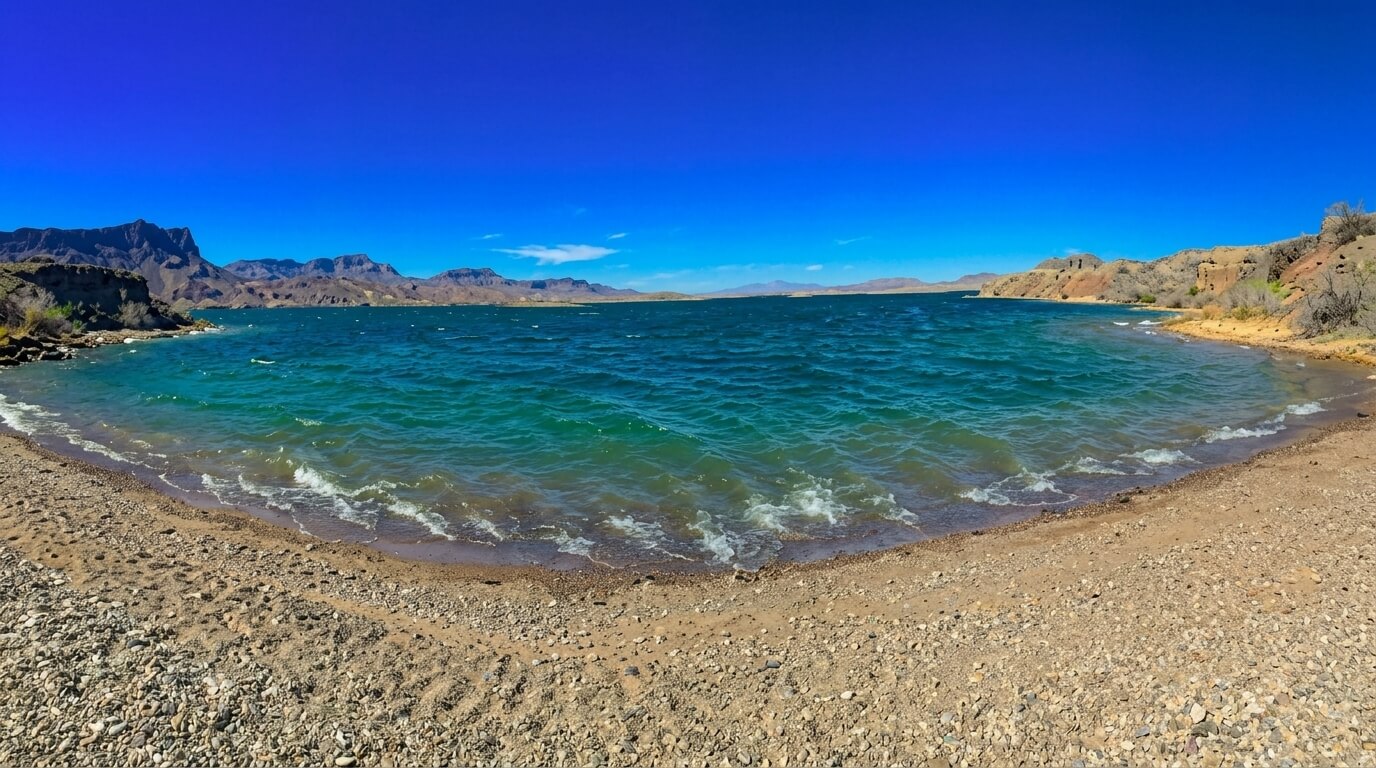 Rocky sandy beach with small waves on a blue lake surrounded by desert mountains under a clear sky