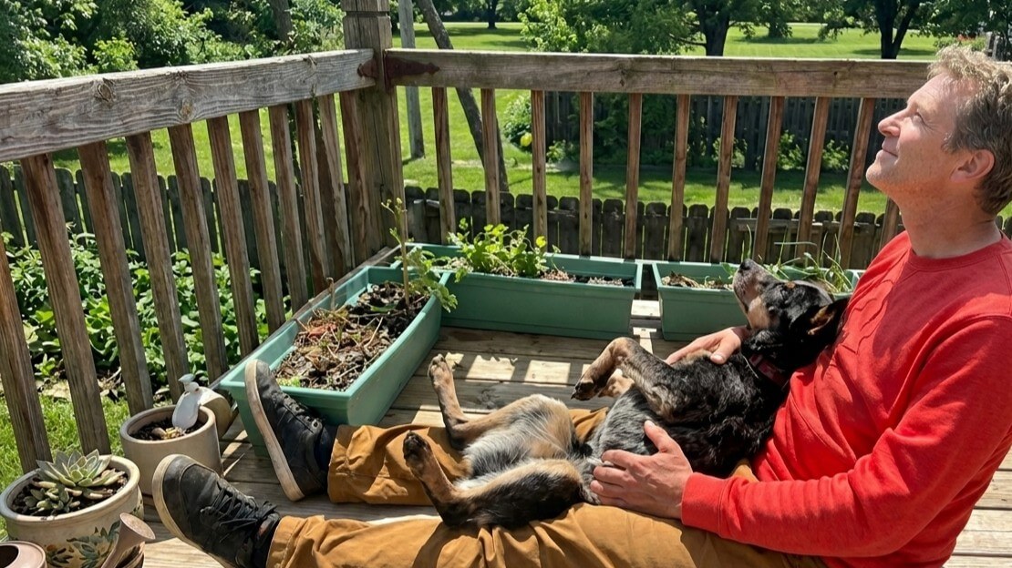 Man in red shirt sitting on wooden deck holding a relaxed black and brown dog on his lap surrounded by potted plants.