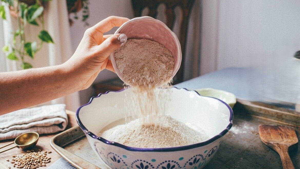 Hand pouring flour from a pink bowl into a large white mixing bowl with blue patterns on a kitchen counter.