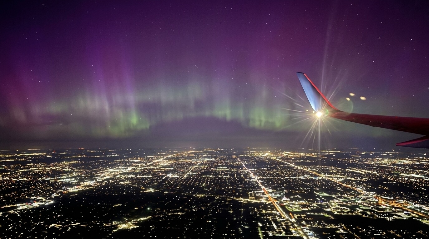 Northern lights over a city at night viewed from an airplane window with the wing and wingtip light visible.