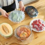 Person covering bowls of food, cantaloupe, and tomatoes with plastic wrap on a wooden table with spices and herbs nearby