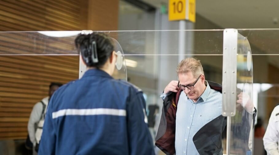 Man removing jacket at airport security checkpoint with officer in blue uniform nearby