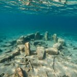 Underwater ancient stone circle ruins on sandy seabed with scattered artifacts and fish swimming nearby