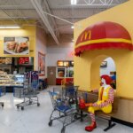 Ronald McDonald statue sitting on a bench under a red and yellow McDonald's awning inside a grocery store.