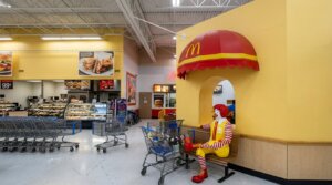 Ronald McDonald statue sitting on a bench under a red and yellow McDonald's awning inside a grocery store.