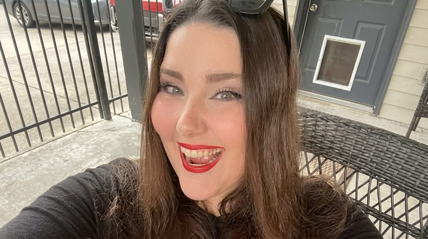Smiling woman with long brown hair, red lipstick, and sunglasses on her head sitting on a wicker chair outdoors.