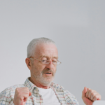 Elderly man with glasses and plaid shirt raising fists in front of a laptop.