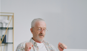 Elderly man with glasses and plaid shirt raising fists in front of a laptop.