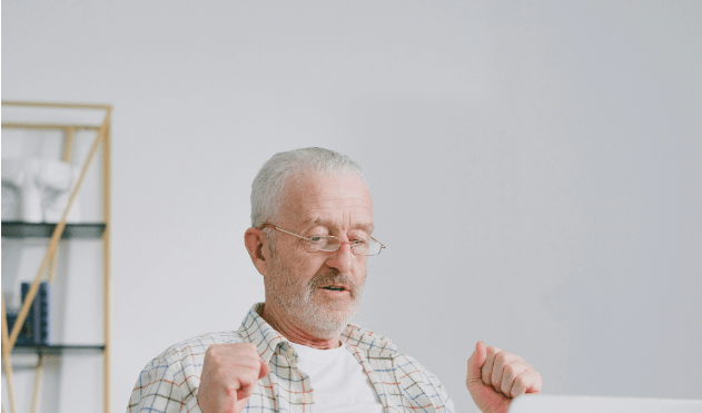 Elderly man with glasses and plaid shirt raising fists in front of a laptop.