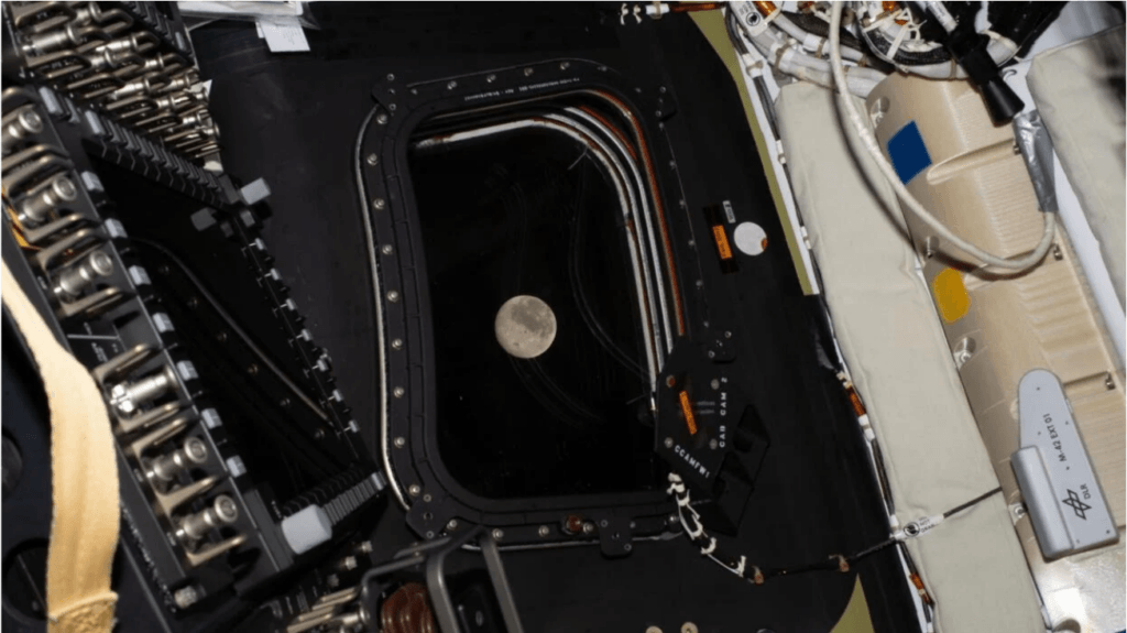 Full moon visible through a spacecraft window surrounded by control panels and wiring inside the cabin