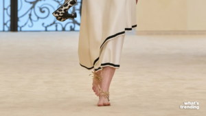 Close-up of a model walking barefoot with gold ankle ties and holding leopard print high heels on a runway.