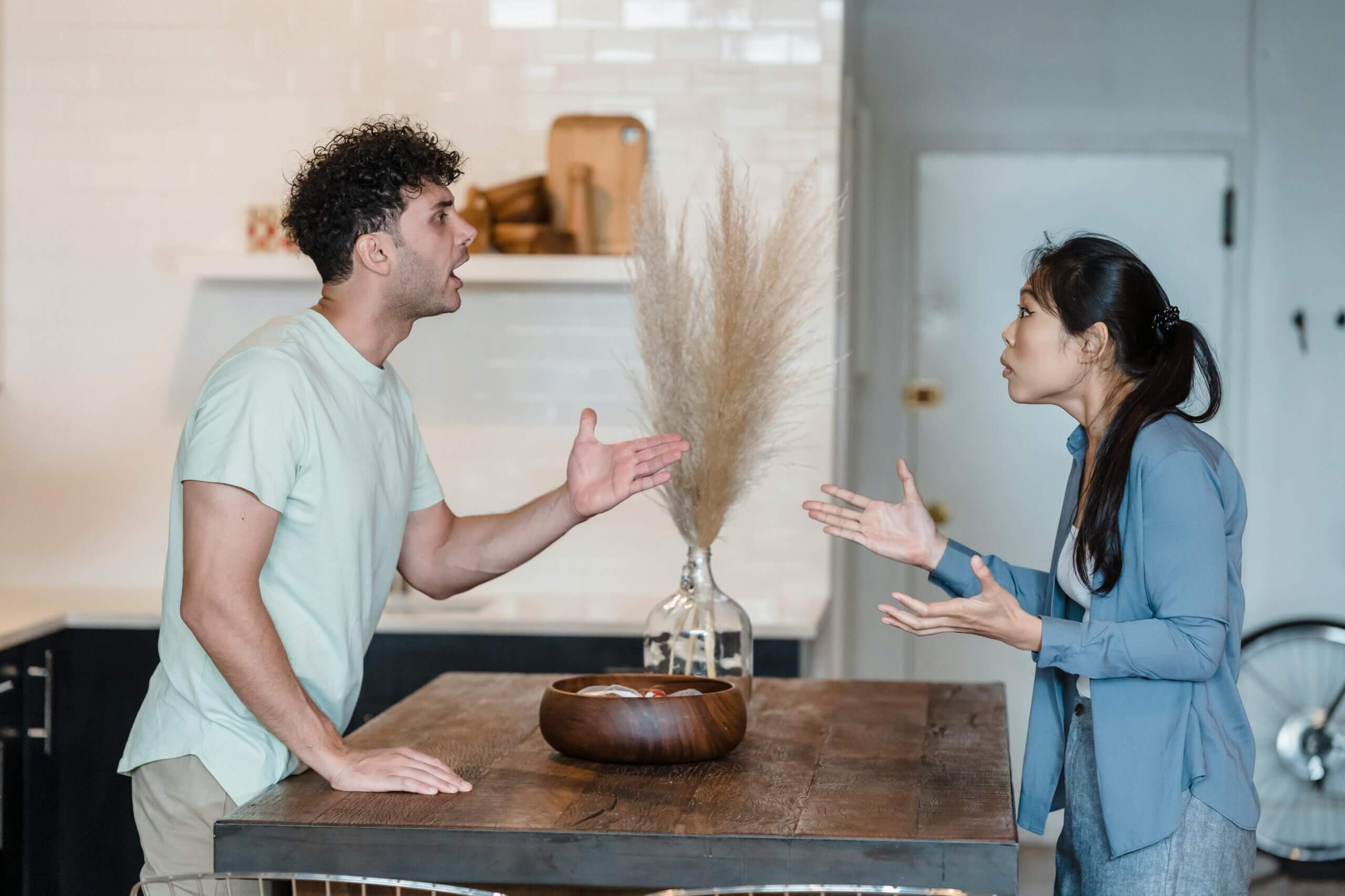 Man and woman arguing across a wooden table in a modern kitchen with dried pampas grass centerpiece