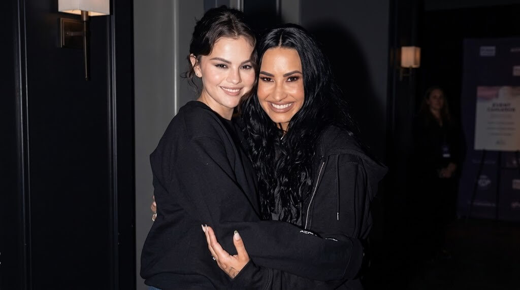 Selena Gomez and Demi Lovato smiling and hugging while wearing black outfits in a dimly lit room.