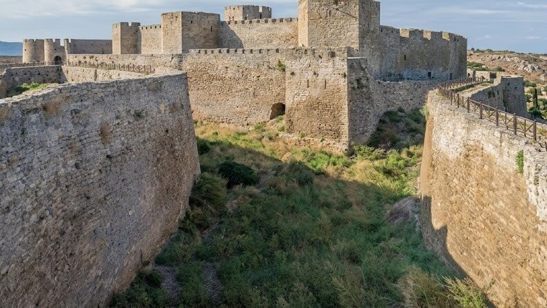 Medieval stone fortress with high walls and grassy moat under a blue sky
