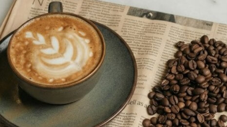 Latte with leaf-shaped latte art in a gray cup and saucer next to a pile of coffee beans on newspaper.