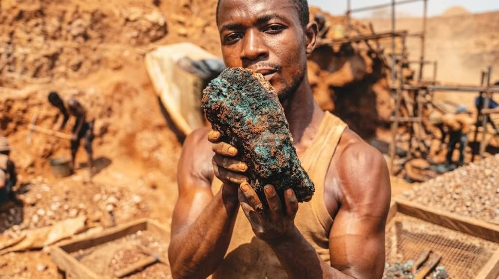 Miner holding a large raw copper ore at a mining site with workers and equipment in the background