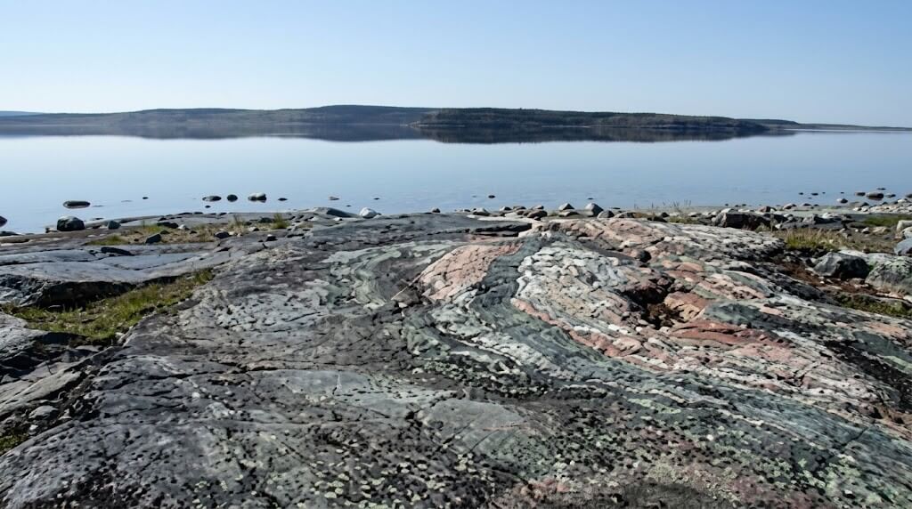 Colorful layered rock formations by a calm lake with a distant forested shoreline under a clear sky.