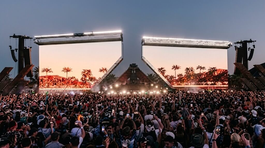 Large crowd at an outdoor music festival with two large screens displaying palm trees and desert scenery at sunset.